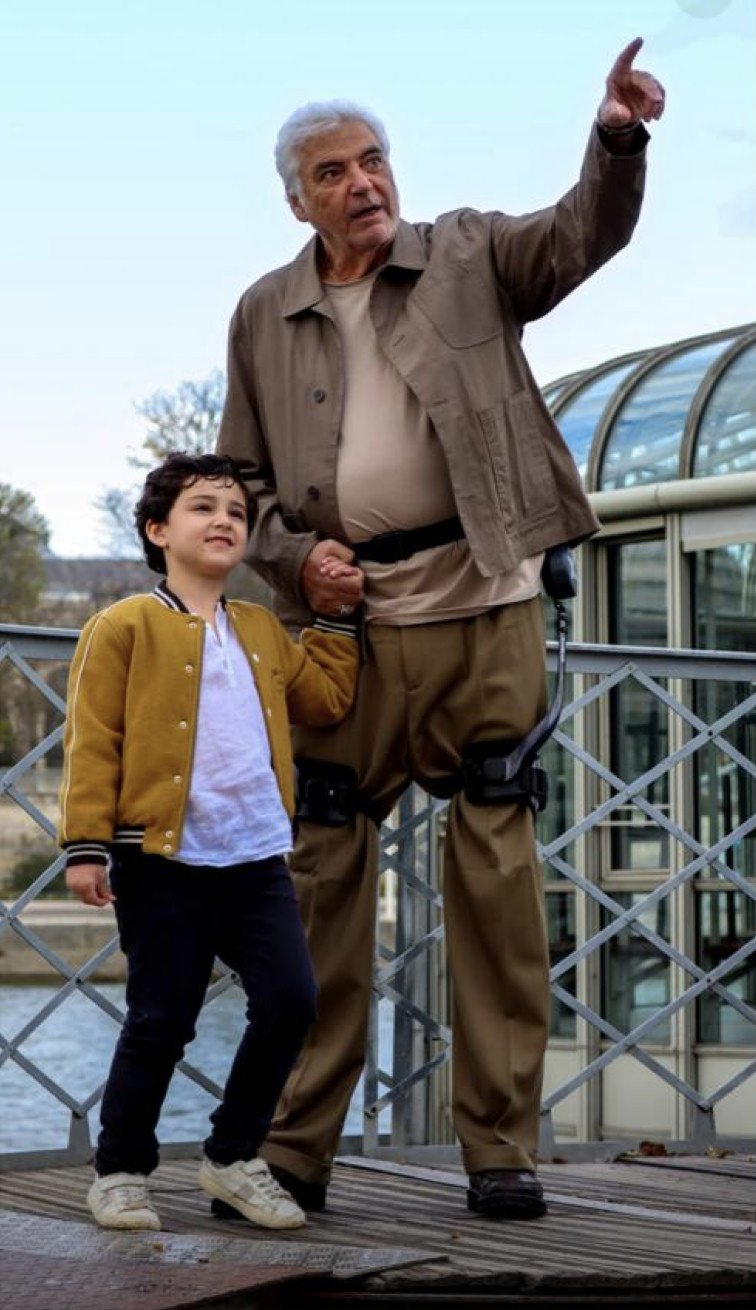 Senior portant l’exosquelette ExoBlue V-X1, main dans la main avec un enfant sur une passerelle de la Seine